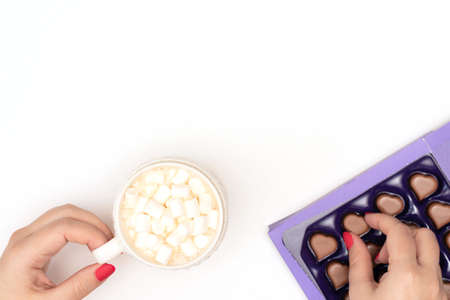 Female hands with a cup of cappuccino with white small marshmallows and a box of heart shaped chocolates on white background. Valentines day, love concept. Flat lay style with copy spaceの写真素材