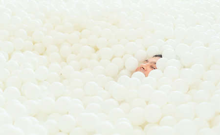 Male face peeking out of the big dry pool with thousand of white balls. Childhood concept. Indoor leisure activity. Having fun in playroomの写真素材