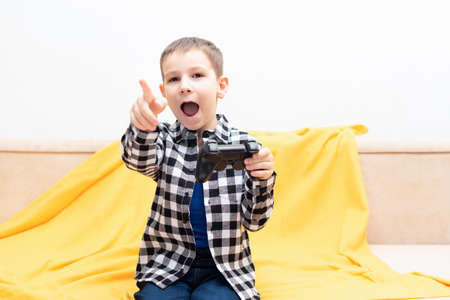 The child boy in checked shirt sitting on the couch with black joystick in his hands emotionally...