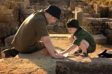 Two archeologists, child and adult, in khaki clothes sitting on ruins and studying an ancient map....