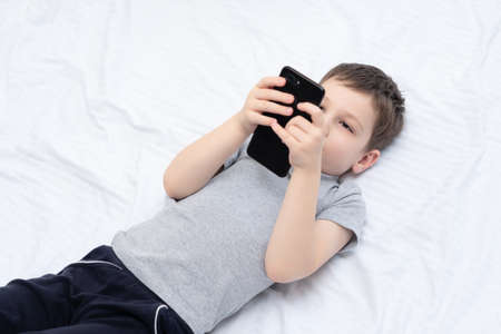Child boy lying on the bed with smartphone playing games or studying something on it. Social and...