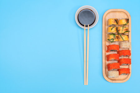Delicious Japanese rolls with salmon, avocado, cucumber, cheese on wooden plate with wood sticks and soy sauce with selective focus on blue background. Asian food concept. Healthy eating.の写真素材