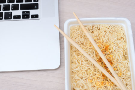 Instant Chinese curly noodles in takeout packaging with wood sticks and laptop on office table. Selective focus. Asian food and delivery concept. Unhealthy eating.の写真素材
