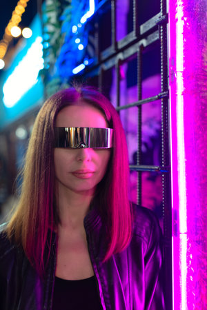 Defocused beautiful young woman in cosmic glasses standing next to colorful neon light at night with selective focus. Party disco neon nightclub vibes.の写真素材