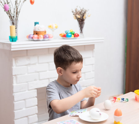 Child boy decorating and painting easter eggs against defocused fireplace with Easter decorations on it. Selective focus. Happy Easter concept. Process of crafting at home. High quality photoの写真素材