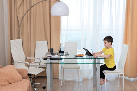 School boy in yellow t-shirt sitting at the table with digital tablet studying at home. Distance...