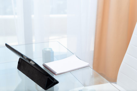 Digital tablet and notebook on glass table with empty white chair prepared for studying at home. Distance learning online education concept. Selective focus. High quality photoの写真素材
