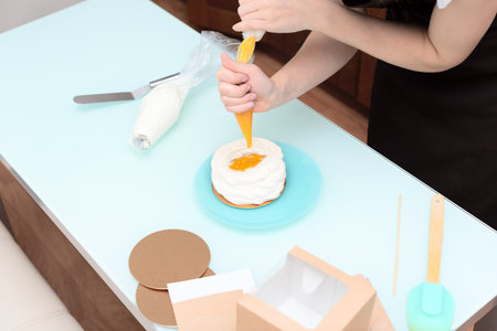 Young woman pastry chef fills mango curd with pastry bag on homemade meringue. Process of making cake Anna Pavlova at home with selective focus. Homemade pastry, delicious sweets concept. High quality photoの写真素材