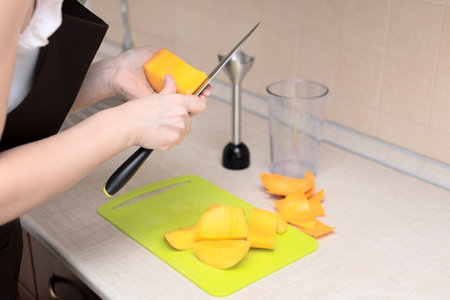 Female chef cutting fresh mango for making homemade tasty mango smoothie or juice. Selective focus. Process of making summer drink at home, healthy lifestyle. High quality photoの写真素材