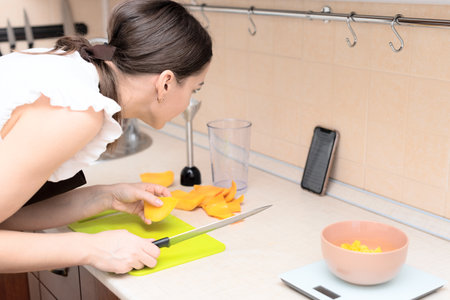 Female chef cutting fresh mango for making homemade tasty mango smoothie or juice using online recipe from smartphone. Selective focus. Process of making summer drink at home, healthy lifestyle. High quality photoの写真素材