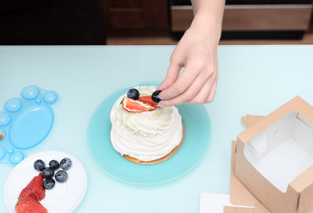 Woman pastry chef at home making finishing touches for homemade meringue cake Pavlova with summer fresh berries with selective focus. Homemade pastry, delicious sweets concept. High quality photoの写真素材