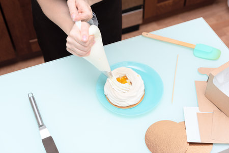 Young woman pastry chef fills cream with pastry bag on homemade meringue. Process of making cake Anna Pavlova at home with selective focus. Homemade pastry, delicious sweets concept. High quality photoの写真素材