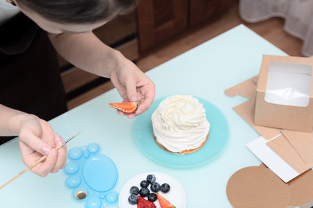 Woman pastry chef at home decorating homemade meringue cake Pavlova with summer fresh berries with selective focus. Homemade pastry, delicious sweets concept. High quality photoの写真素材