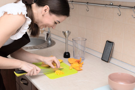 Female chef cutting fresh mango for making homemade tasty mango smoothie or juice using online recipe from smartphone. Selective focus. Process of making summer drink at home, healthy lifestyle. High quality photoの写真素材
