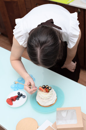Woman pastry chef at home making finishing touches for homemade meringue cake Pavlova with summer fresh berries with selective focus. Homemade pastry, delicious sweets concept. High quality photoの写真素材