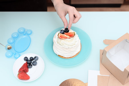 Woman pastry chef at home making finishing touches for homemade meringue cake Pavlova with summer fresh berries with selective focus. Homemade pastry, delicious sweets concept. High quality photoの写真素材