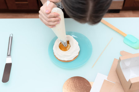 Young woman pastry chef fills cream with pastry bag on homemade meringue. Process of making cake Anna Pavlova at home with selective focus. Homemade pastry, delicious sweets concept. High quality photoの写真素材