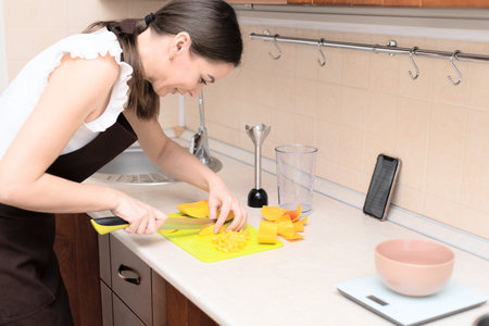 Female chef cutting fresh mango for making homemade tasty mango smoothie or juice using online recipe from smartphone. Selective focus. Process of making summer drink at home, healthy lifestyle. High quality photoの写真素材