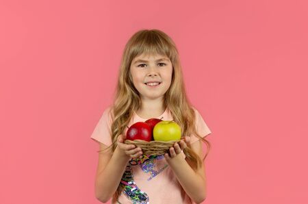 Beautiful little girl on a pink background holding a basket with apples. Girl with apples in her hands. Healthy food concept. Basket with apples. Little girl on a pink background. Harvest season.の写真素材