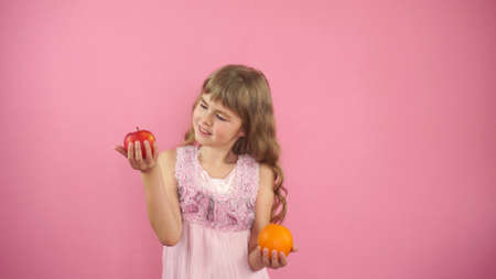 Little girl on a pink background holding an apple and an orange. The child makes a choice between an...