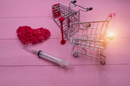 In selective focus of the syringe and woven red heart put beside blurred shopping cart,on pink pastel timber board,warm light tone,blurry light design backgroundの写真素材