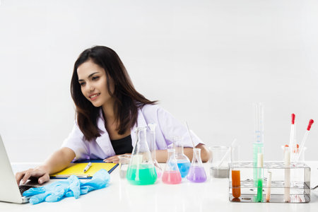 The chemist is working by test tube with reagent in lab,she is using laptop for searching data with...