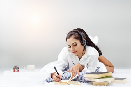 Lady in white shirt is laying down on bed,writing on book,listening music,relax time,blurry light aroundの写真素材