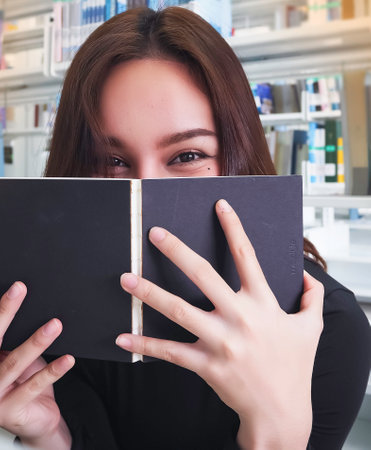 Young beautiful lady put book cover her face and smile in eyes,in front of blurred bookshelf.blurry...