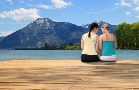 Two young woman sitting on a wooden deck with their backs to the camera chatting overlooking a picturesque lake with mountain backdropの写真素材