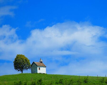 Quaint little chapel with a tiny tower under a solitary tree on the skyiline of a lush green hilltopの写真素材
