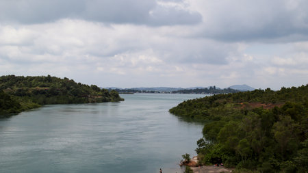A large river with green trees on the left and right ended into a lake with a mountain background in the horizon with bright cloudy sky.の写真素材