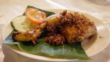 Chicken Serudeng or Fried Chicken with Fried Coconut Flakes, some cucumber and tomato sliced served on top of banana leaf in a plate. Indonesian Food.の写真素材