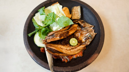 Crispy fried milkfish served with spicy sambal penyet and fresh vegetables on a rustic black plate in Indonesia featuring vibrant garnishes and traditional presentation.の写真素材