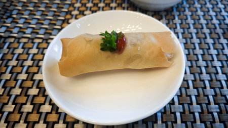 Crispy Spring Roll Garnished with Parsley and Chili Sauce on White Plate with Woven Table Mat Backgroundの写真素材