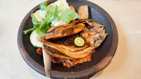 Crispy fried milkfish served with spicy sambal penyet and fresh vegetables on a rustic black plate in Indonesia featuring vibrant garnishes and traditional presentation.の写真素材