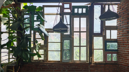 Rustic indoor space with vintage mismatched window frames, hanging industrial lamps, green leafy plants, and exposed brick walls letting in natural daylight with a garden view outsideの写真素材