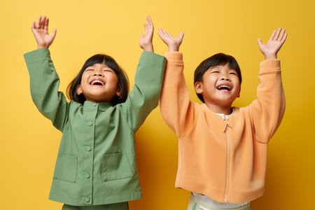 Portrait of two little Asian kids raising hands up isolated over yellow backgroundの素材