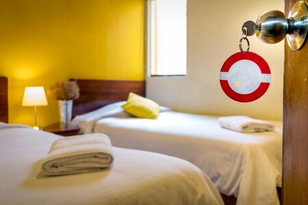 Hotel room with the key ring to complete. In the background you can see two beds, a small vertical window, towels, a vase and a lampの写真素材