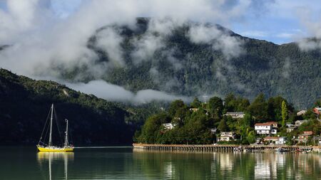 Beautiful panoramic view of the city of Caleta Tortel in Patagonia Chile. There are several houses between the forest that are reflected in the Pacific Ocean and a yellow sailboat in the water.の写真素材