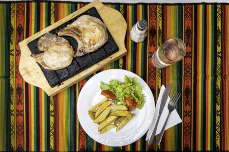 Plate of french fries with tomato and lettuce salad. A wooden tray containing a stone that keeps warm meat from sifting. Colorful tablecloth, glass with water, salt shaker and cutleryの写真素材