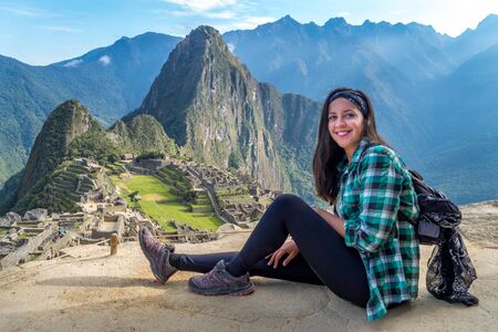 A woman sitting on a Machu Picchu terrace. You can see the ruins of the citadel of Machu Picchu, the Huayna Picchu mountain and other huge mountains behind.の写真素材