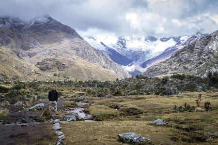 Trail to Laguna 69. A person standing posing. There are small bushes around it and huge mountains behind with snowy peaks and clouds. Peru, white mountain range. Latin Americaの写真素材