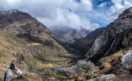 A man sitting on a rock in a mountain landscape in the Cordillera Blanca, Peru. There is a great valley and a waterfall. Mount Huascaran with a background full of cloudsの写真素材