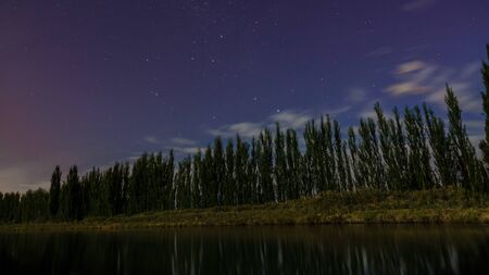 Night photography at a river with a line of trees is reflected on the water. Some stars and clouds in the sky Blue hour. Patagonia, Argentinaの写真素材
