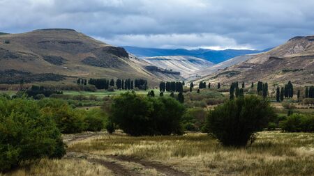 Valley of green and fresh grass, many bushes and a beautiful tree to the left of the photograph. In the background you can see the mountains, poplars and a sky covered with clouds. Argentina, Patagoniaの写真素材