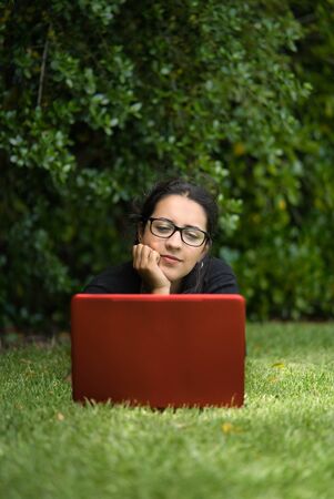 Woman lying down on the grass of a park looking at the screen her laptop. She seems to be thinking about something. Natural Environment. Vertical Photographyの写真素材