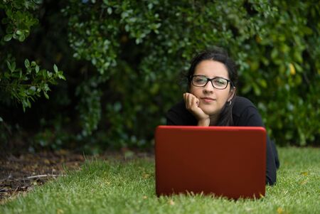 Brunette woman lying down on the grass with her computer. She's is looking at the camera. Natural Environment. Technological conceptの写真素材