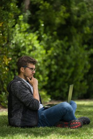 A young blond man is sitting on the grass of a park with his laptop. He is with her legs crossed and he seems to be thinking about something. Natural environment. Vertical photographyの写真素材