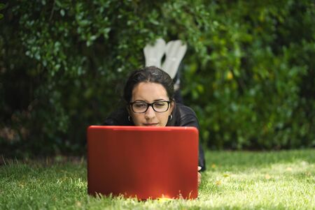 Woman lying down on the grass of a park working on her laptop with her feet up. Natural Environment