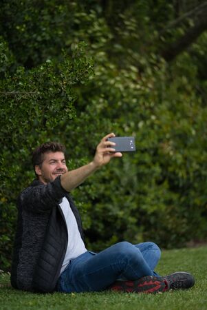 Blond young man sitting on the grass of a park and taking a selfie with his mobile phone while he is smiling. Technological concept. Vertical photographyの写真素材